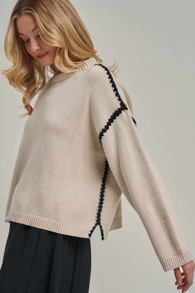A woman wearing a cropped beige knit sweater with black decorative trim along the shoulder and sleeve, paired with dark pleated trousers, against a light gray studio background.