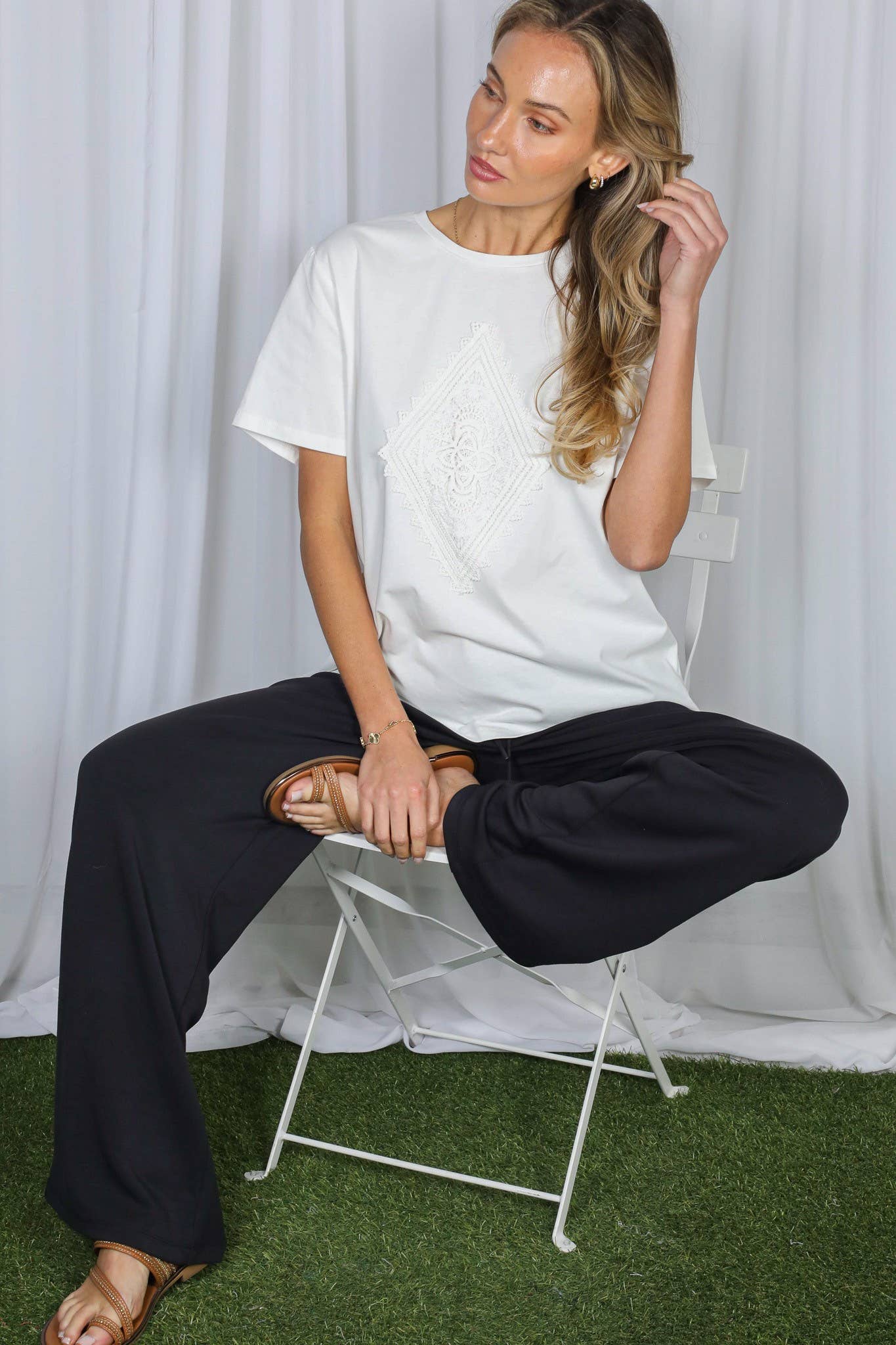A woman sits on a white folding chair wearing a white short-sleeve t-shirt with a subtle tonal graphic and black wide-leg pants, set against a white curtain backdrop with green artificial grass at the bottom.