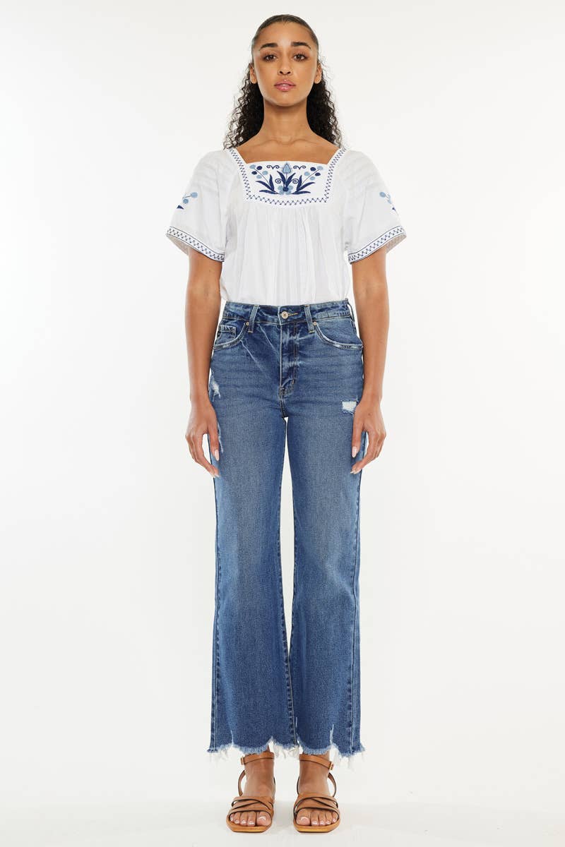 A woman modeling a white short-sleeve top with blue floral embroidery on the chest and sleeves, paired with high-waisted blue wide-leg jeans and brown sandals, against a plain white background.
