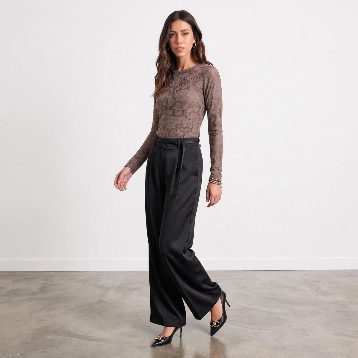 Model wearing Apricot Lane Duluth black satin belted pants with a brown lace long-sleeve top and black heels