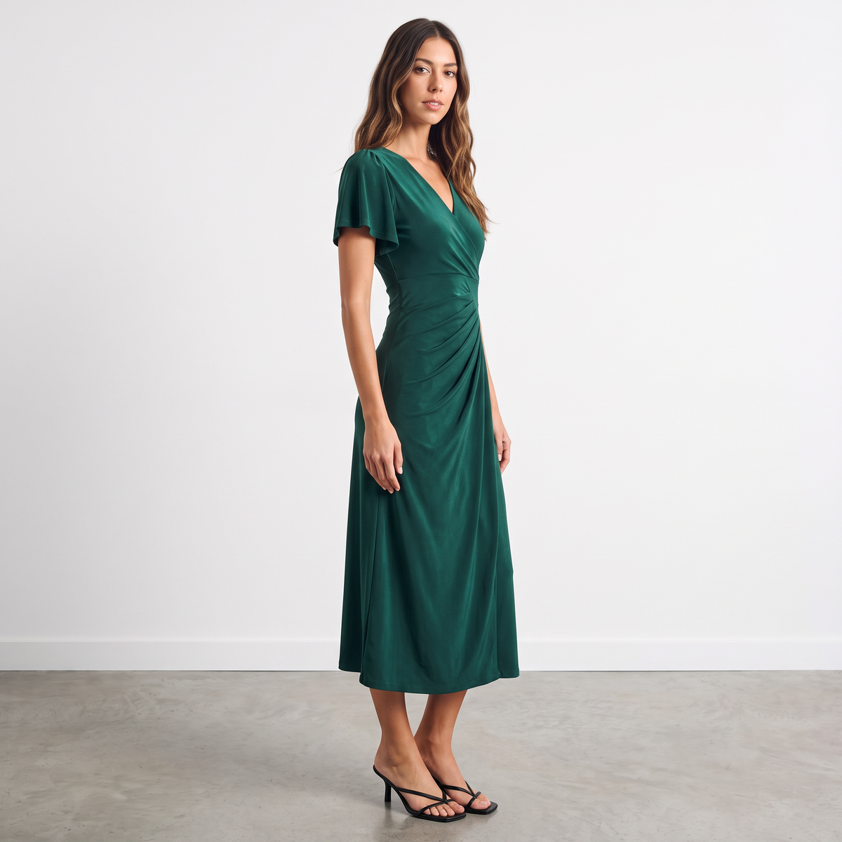 Model wearing an Apricot Lane Duluth emerald green midi dress with flutter sleeves and asymmetric pleated detailing.