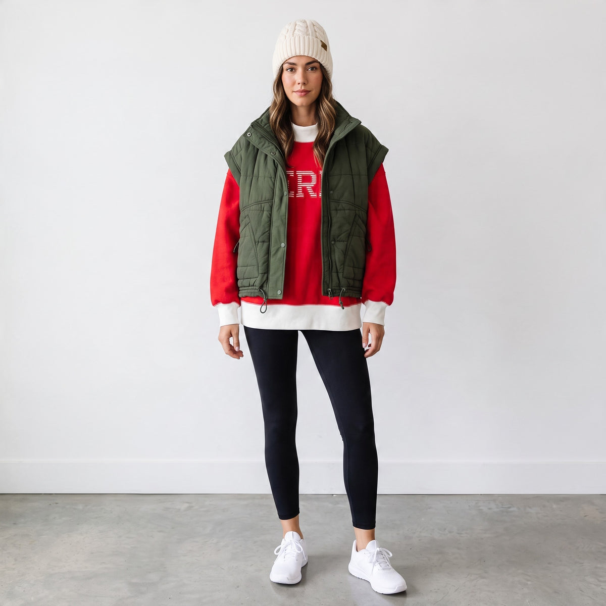 Apricot Lane Duluth model wearing a green puffer vest over a red pullover, black leggings, and a cream beanie