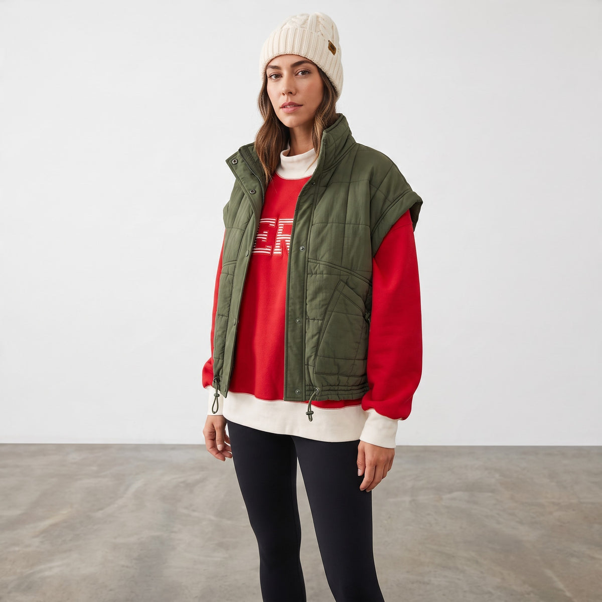 Apricot Lane Duluth model wearing an olive green puffer vest over a red sweatshirt, black leggings, and a beanie.