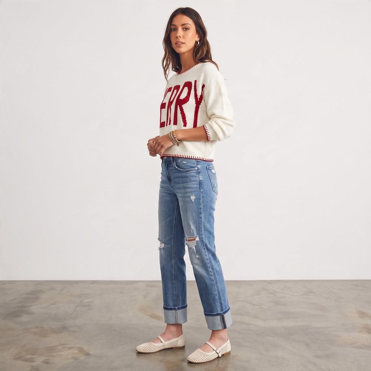 Apricot Lane Duluth cream knit sweater with red MERRY graphic, red trim, and cuffed denim jeans on a female model