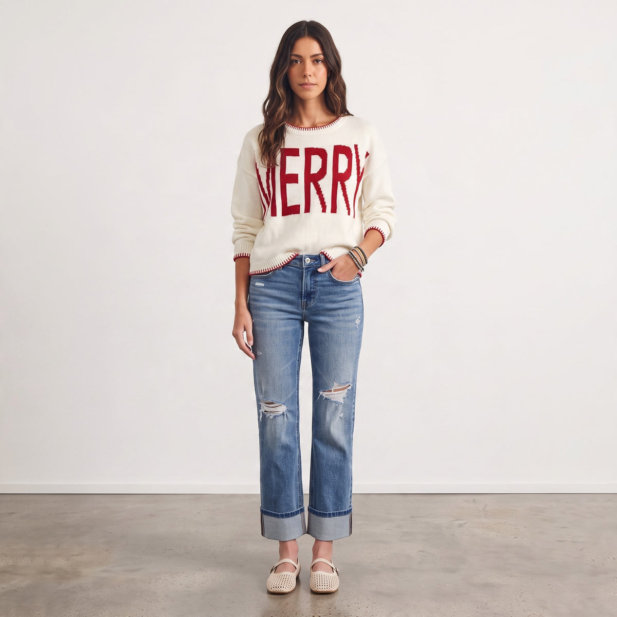 Model wearing an Apricot Lane Duluth cream knit sweater with red MERRY graphic, red trim, and distressed blue jeans.