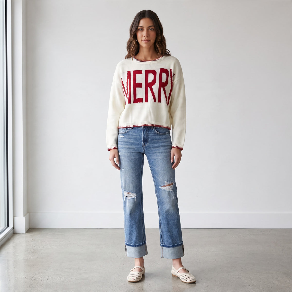 Model wearing an Apricot Lane Duluth cream knit sweater with red MERRY graphic and red trim paired with blue jeans