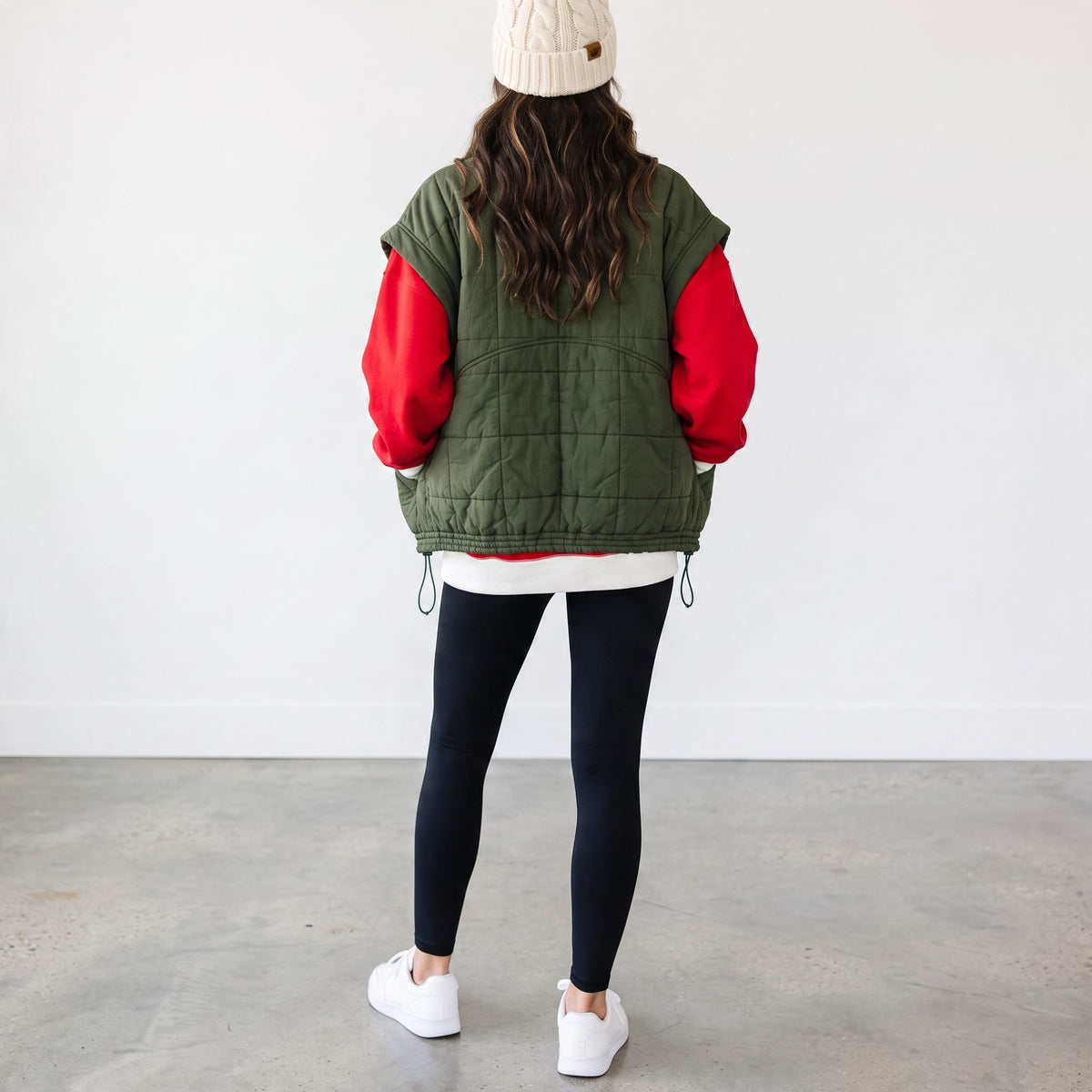 Apricot Lane Duluth olive green quilted puffer vest layered over a red sweatshirt with black leggings and a beanie