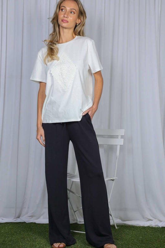 A woman modeling a white short-sleeve t-shirt with a textured, raised circular pattern on the front, paired with dark wide-leg pants, standing in front of a white draped fabric backdrop.