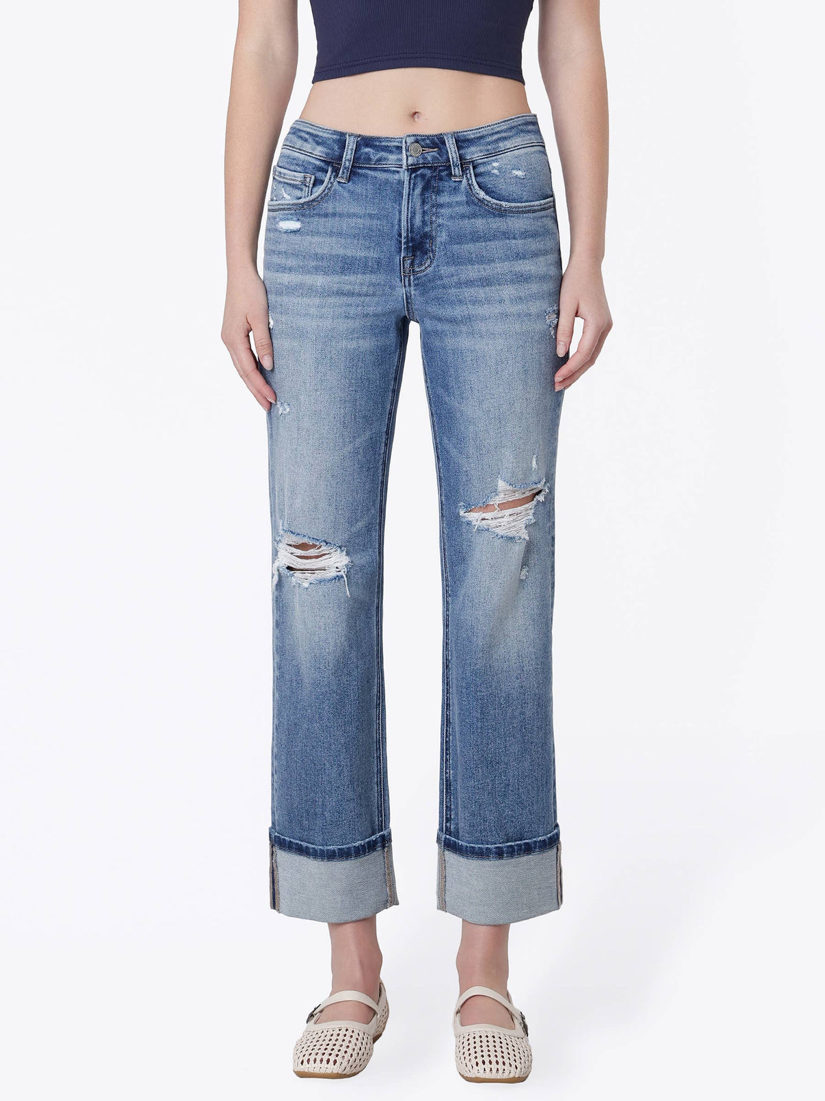 A model wearing a pair of light blue, straight-leg cropped jeans with distressed rips on the thighs and rolled hems, paired with a dark crop top and beige woven sandals.