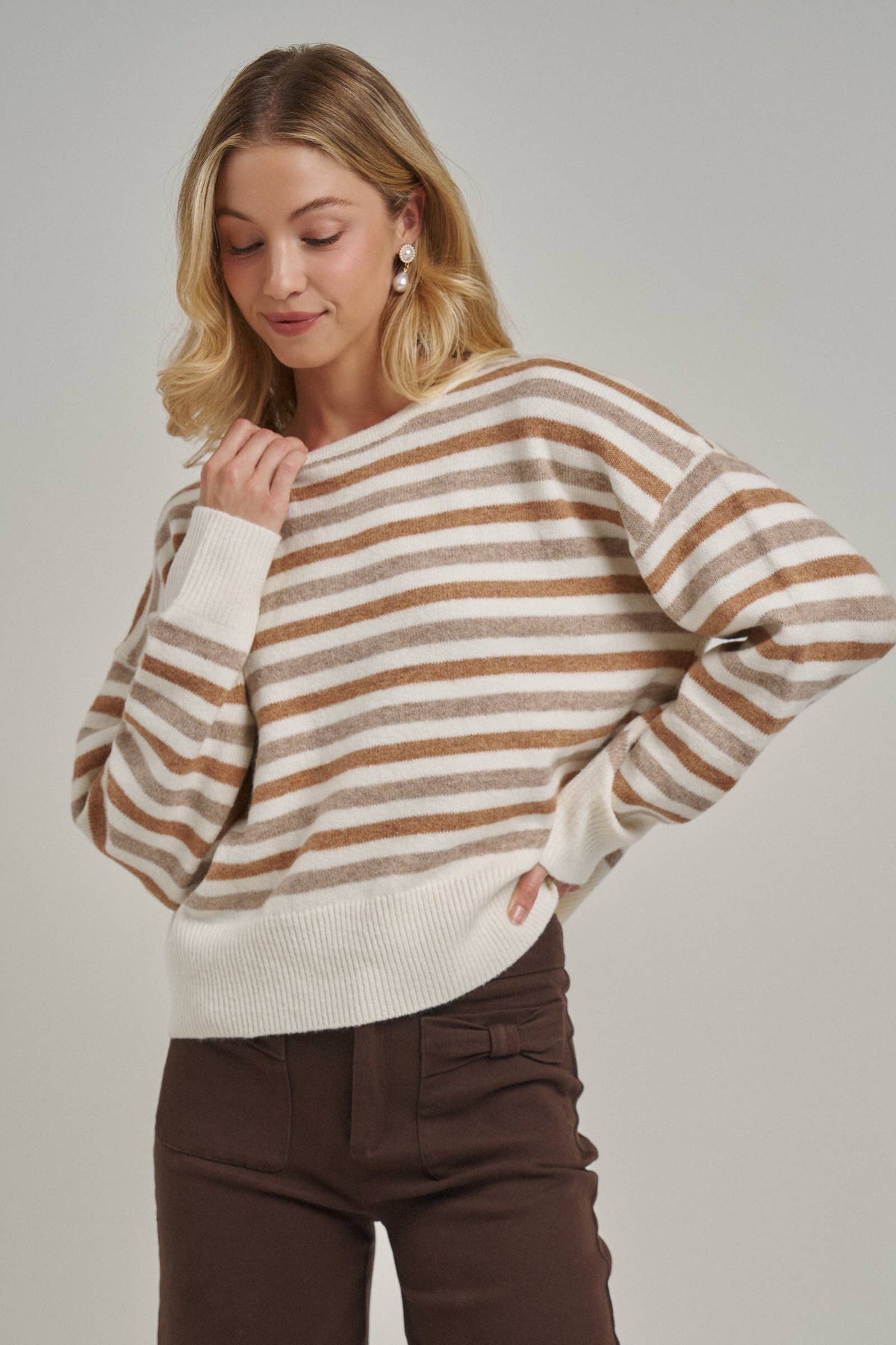 A woman wearing a cream and brown horizontally striped knit sweater with ribbed cuffs and hem, paired with dark brown pants, against a solid light gray studio background.