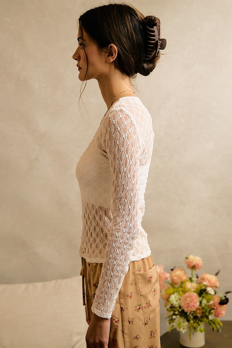 A woman in profile wearing a cream-colored, long-sleeve lace knit top and a beige floral skirt, standing in a softly lit room with a textured beige wall and a vase of pink flowers on a table.
