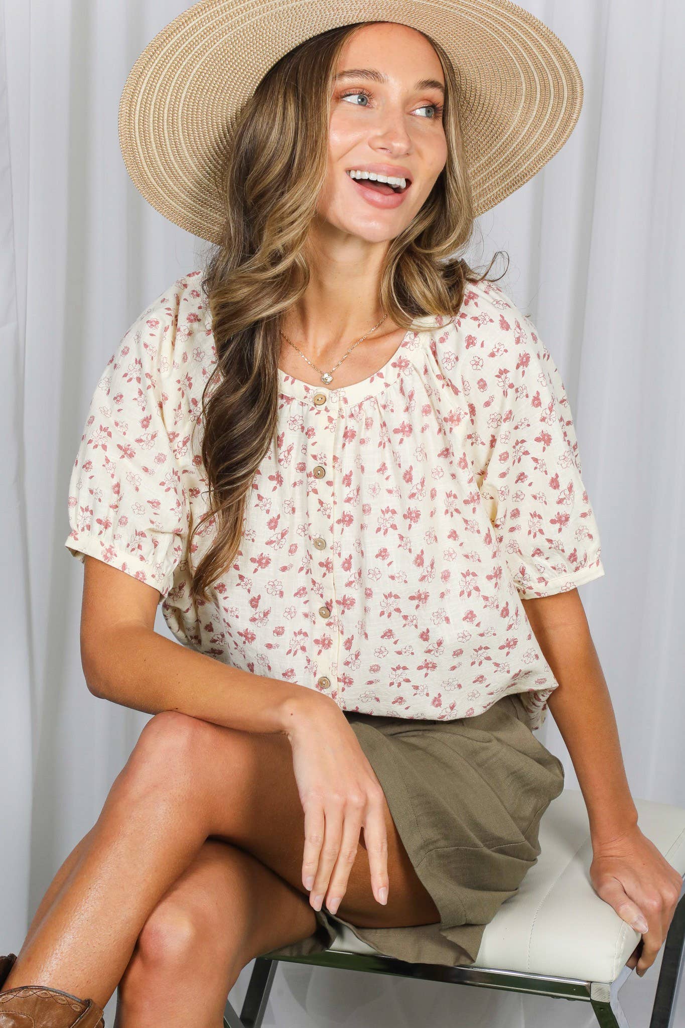 Woman wearing a floral blouse and straw hat sitting on a chair against a white curtain background