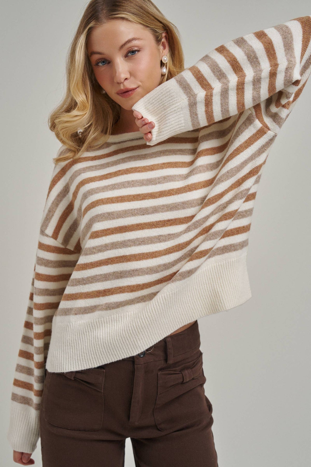 A woman wearing a loose, off-white knit sweater with horizontal tan stripes and dark brown pants, posing against a light gray studio background.