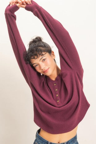 A woman wearing a ribbed knit long-sleeve henley top in a deep plum purple, stretching her arms overhead against a plain off-white background.