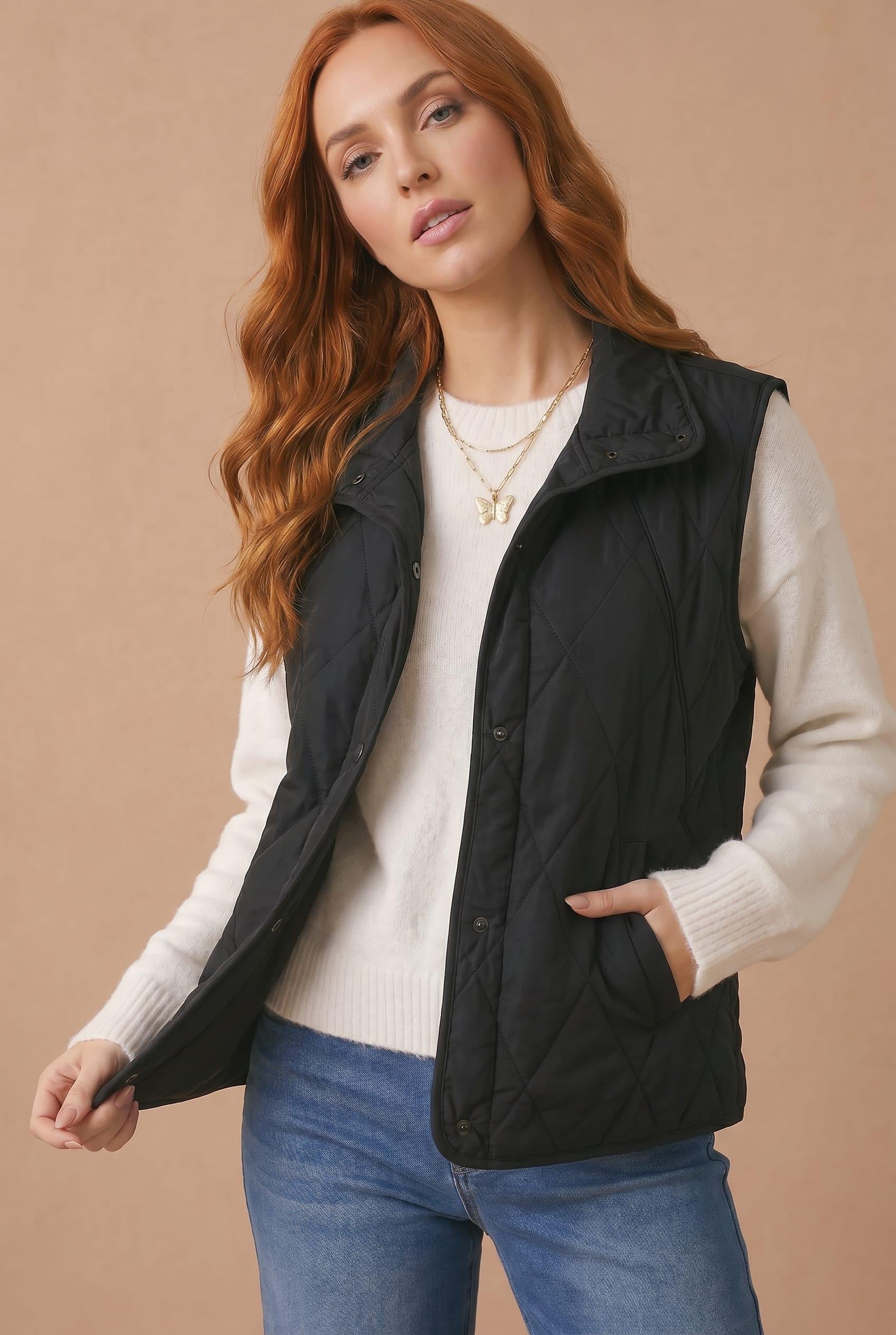 A woman with long wavy red hair models a black quilted sleeveless puffer vest over a cream ribbed knit sweater and blue jeans, set against a solid beige studio background.