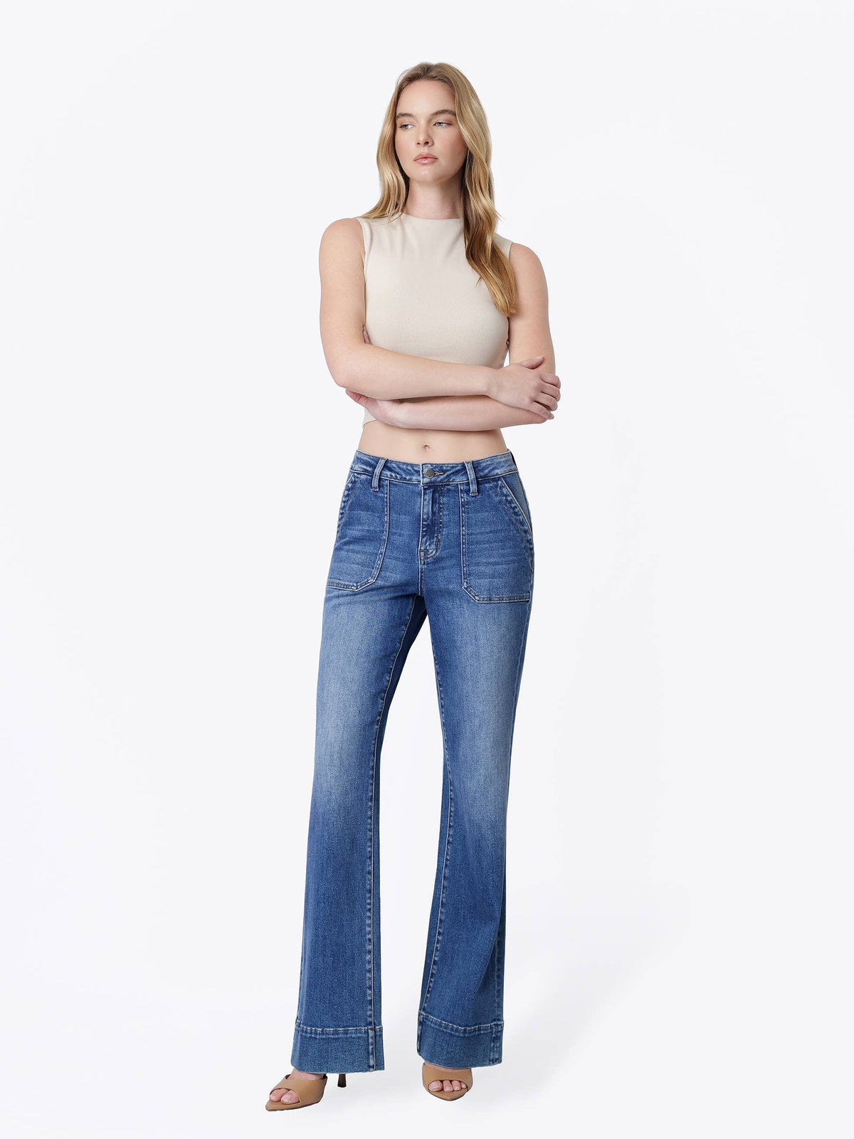 A woman modeling a pair of high-waisted, wide-leg blue denim jeans with a faded wash, paired with a beige sleeveless crop top and nude heels, against a plain white studio background.