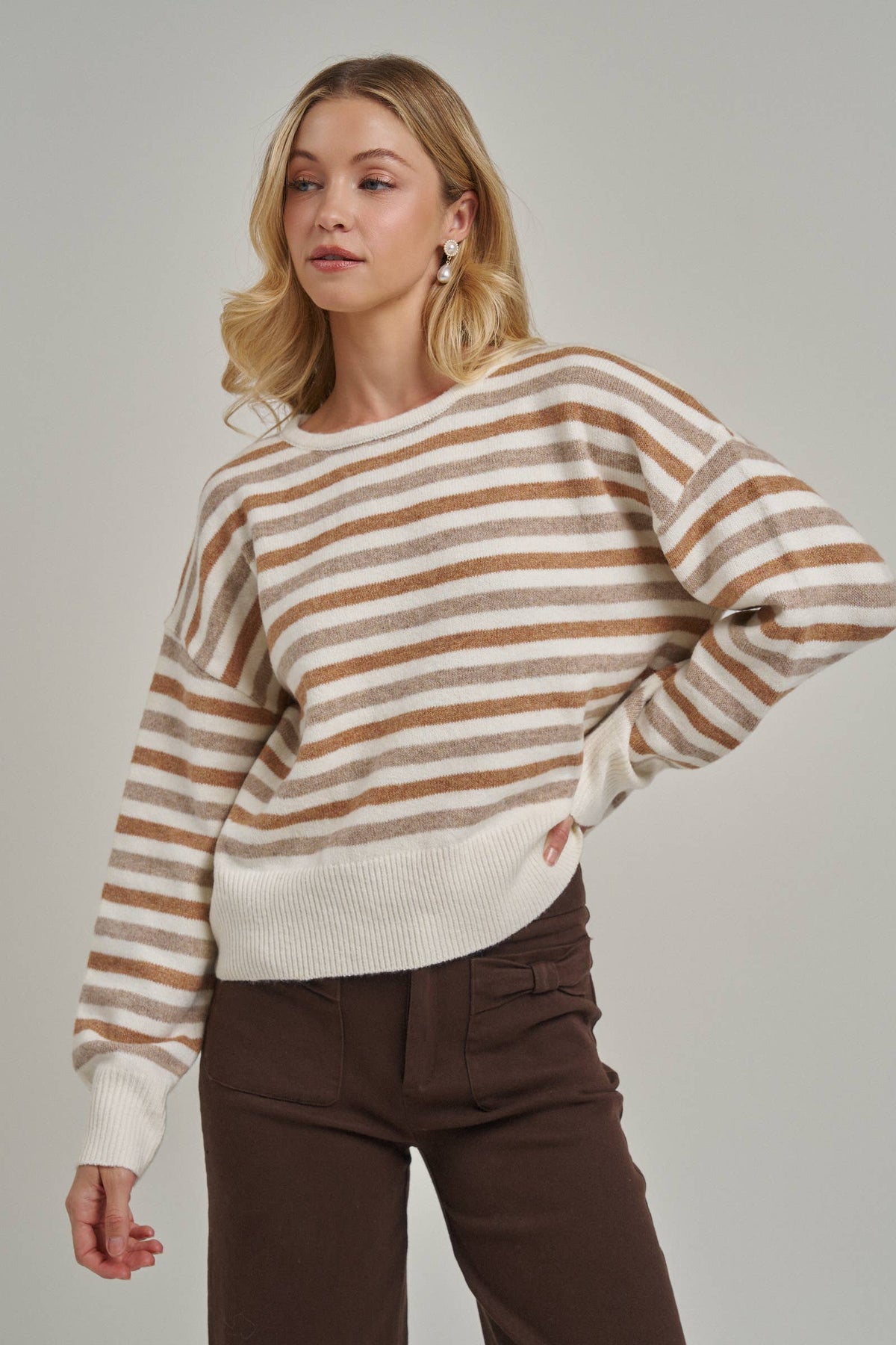 A woman wearing a cream and brown horizontally striped knit sweater with long sleeves, paired with dark brown pants, against a solid light gray studio background.