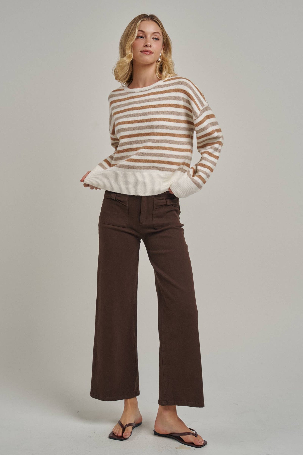 A woman modeling a cream and tan striped knit sweater paired with dark brown wide-leg cropped pants and black flip-flops, standing against a light gray studio background.