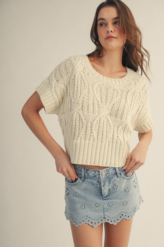 Woman wearing a cream knitted top and denim shorts on a plain background