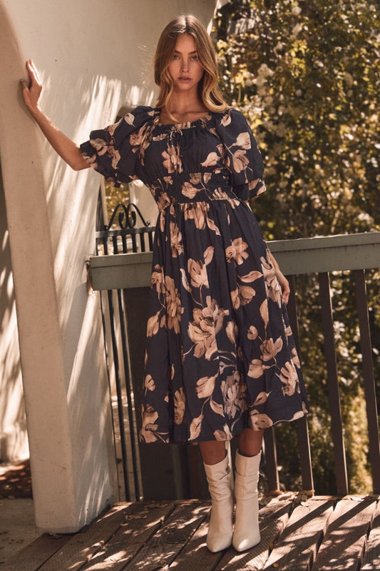Woman in a floral dress standing on a wooden deck with greenery in the background