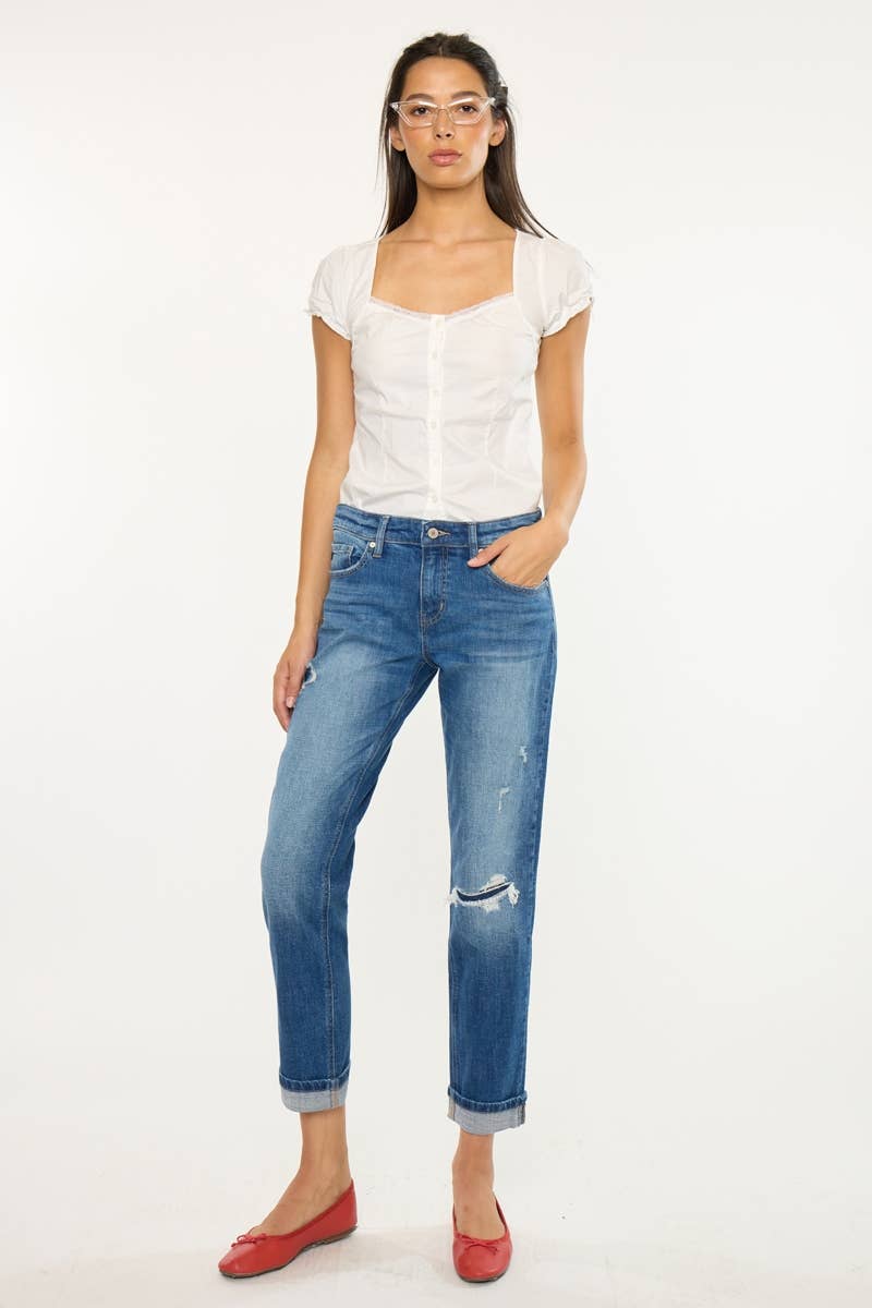 A woman modeling a pair of blue cropped jeans with distressed details and rolled cuffs, paired with a white top and red flats, against a plain white background.