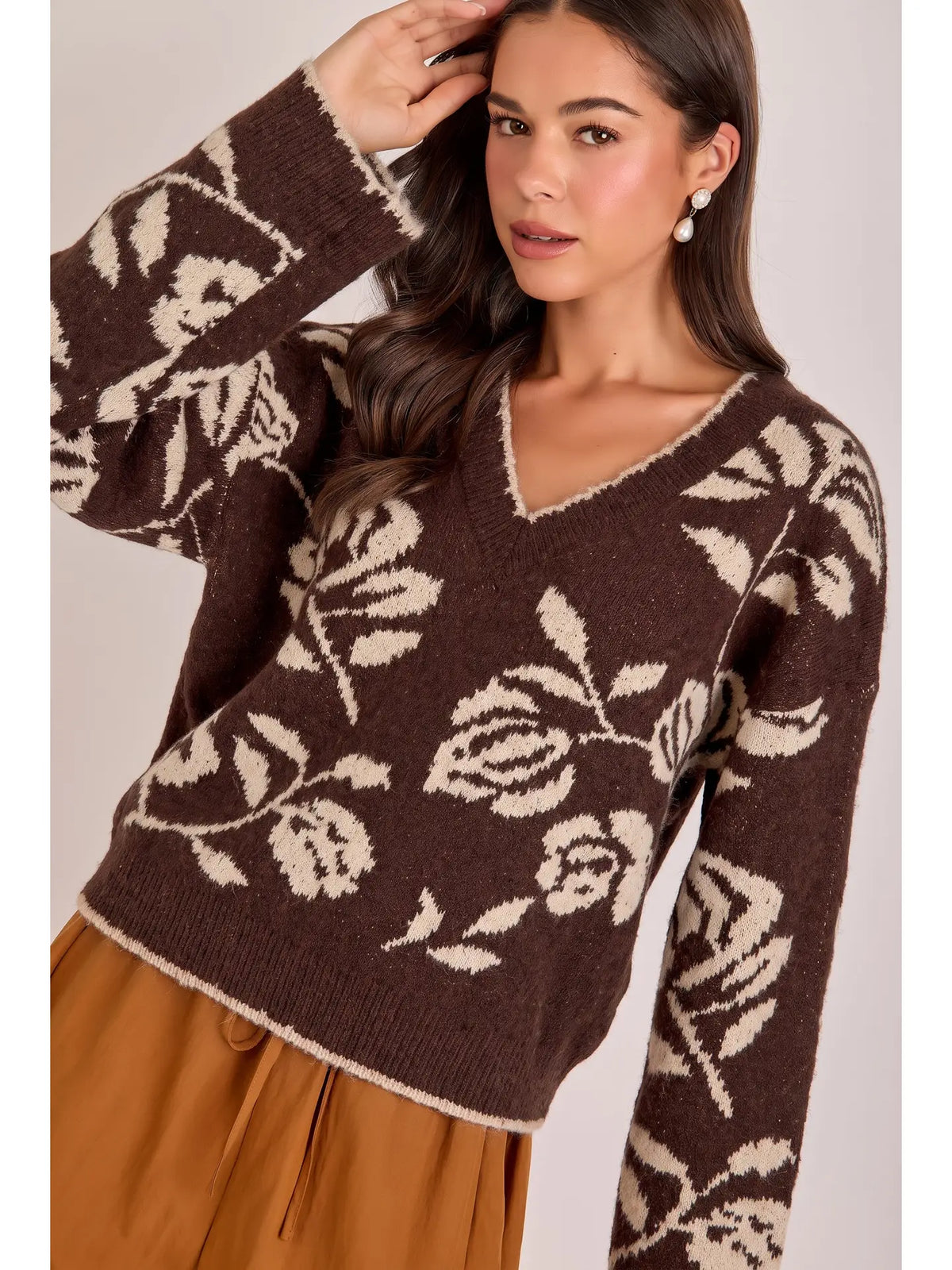 A woman wearing a dark brown knit sweater with a cream floral pattern and a V-neckline, paired with brown pants, against a solid light beige background.