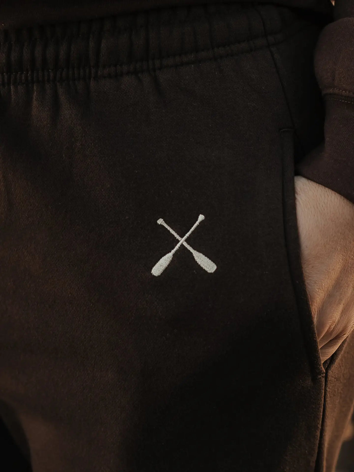 Close-up of dark brown sweatpants with a small white embroidered crossed oars logo on the left hip, shown against a dark background with a hand partially visible in the pocket.