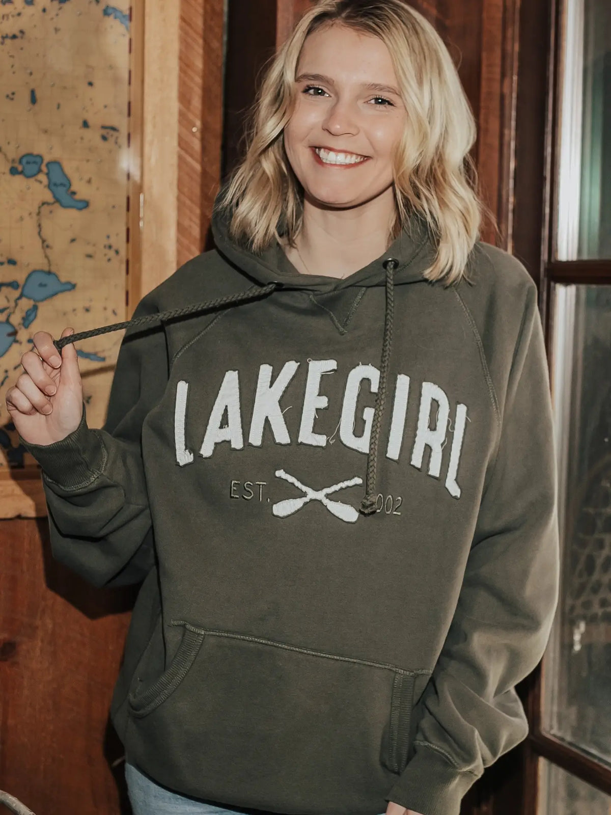 A smiling blonde woman wearing an olive green pullover hoodie with 'LAKEGIRL EST. 2012' and crossed oars printed in white on the front, standing indoors near a wooden door and map.