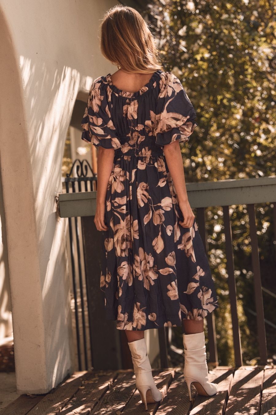 Woman in a floral dress standing on a wooden deck with greenery in the background