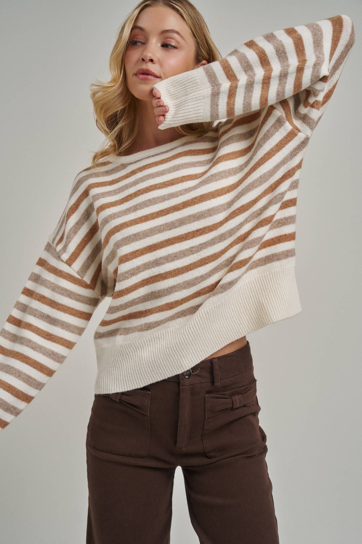 A woman wearing a loose, cropped knit sweater with horizontal brown and cream stripes, paired with dark brown pants, against a solid light gray studio background.
