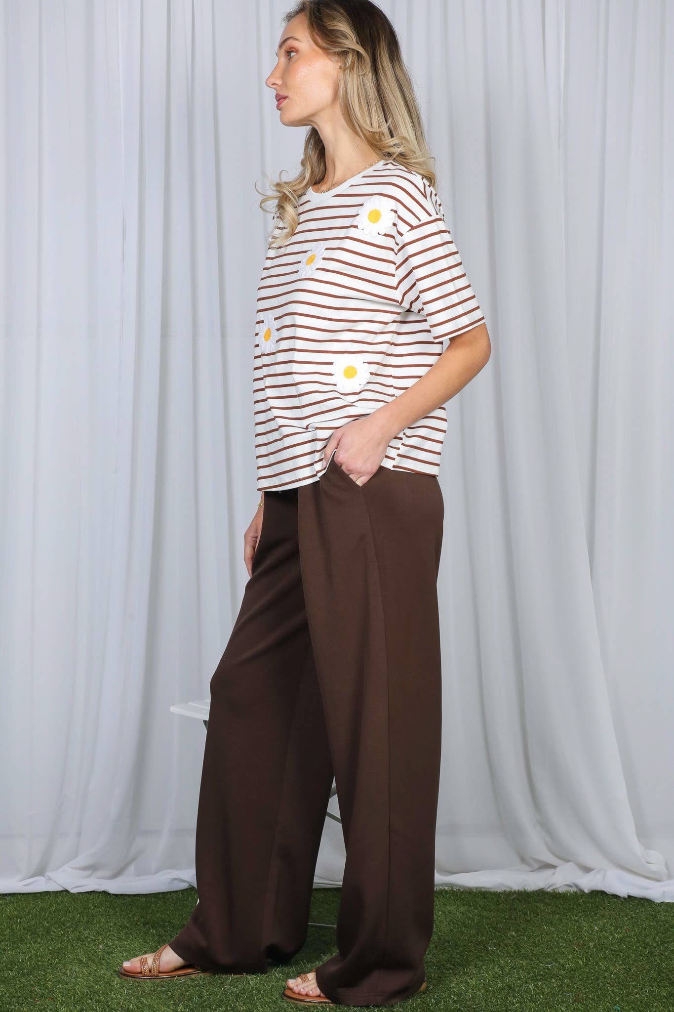 A woman modeling a short-sleeve crewneck t-shirt with thin brown and white horizontal stripes and large white daisy graphics, paired with wide-leg dark brown pants, standing against a white draped curtain backdrop.