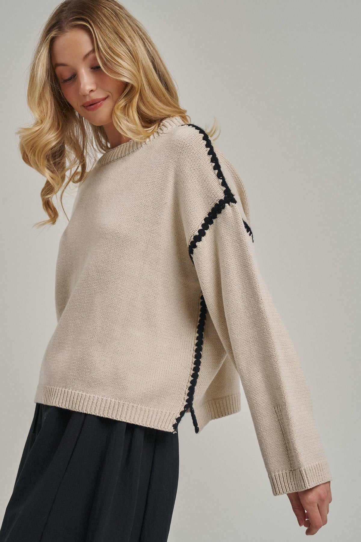 A woman wearing a cream-colored knit sweater with black braided trim details along the shoulder and sleeve, paired with dark pleated trousers, photographed against a neutral light gray background.