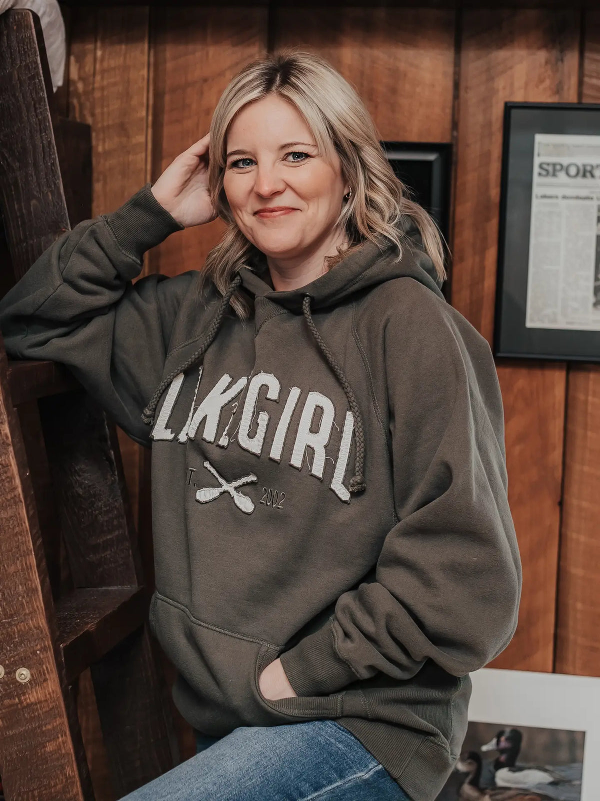 A woman with blonde hair wearing a dark olive green hoodie with 'LKGIRL' printed in white across the chest, posing in front of a rustic wooden wall.