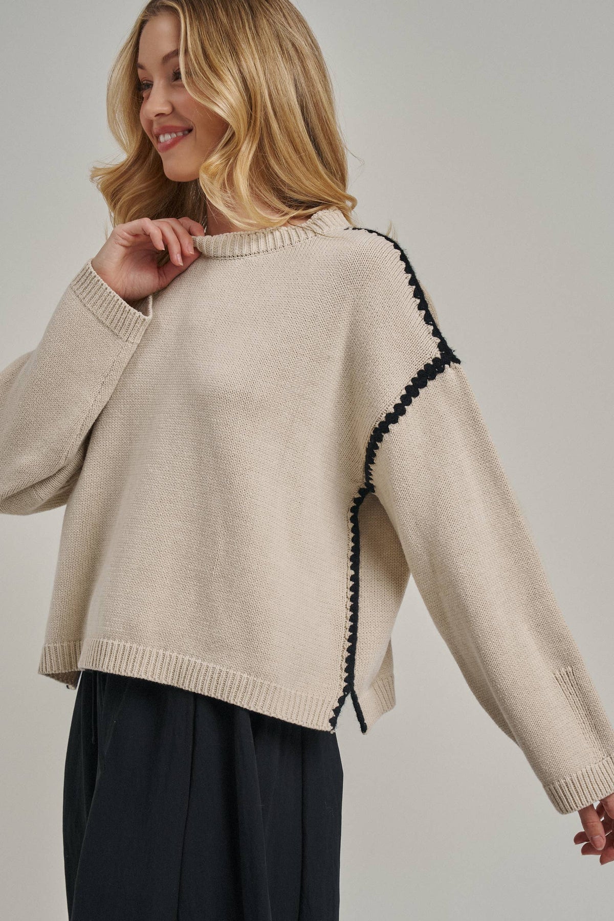 A woman wearing a cream-colored knit sweater with black decorative trim along the shoulder seam, paired with dark trousers, against a neutral studio background.