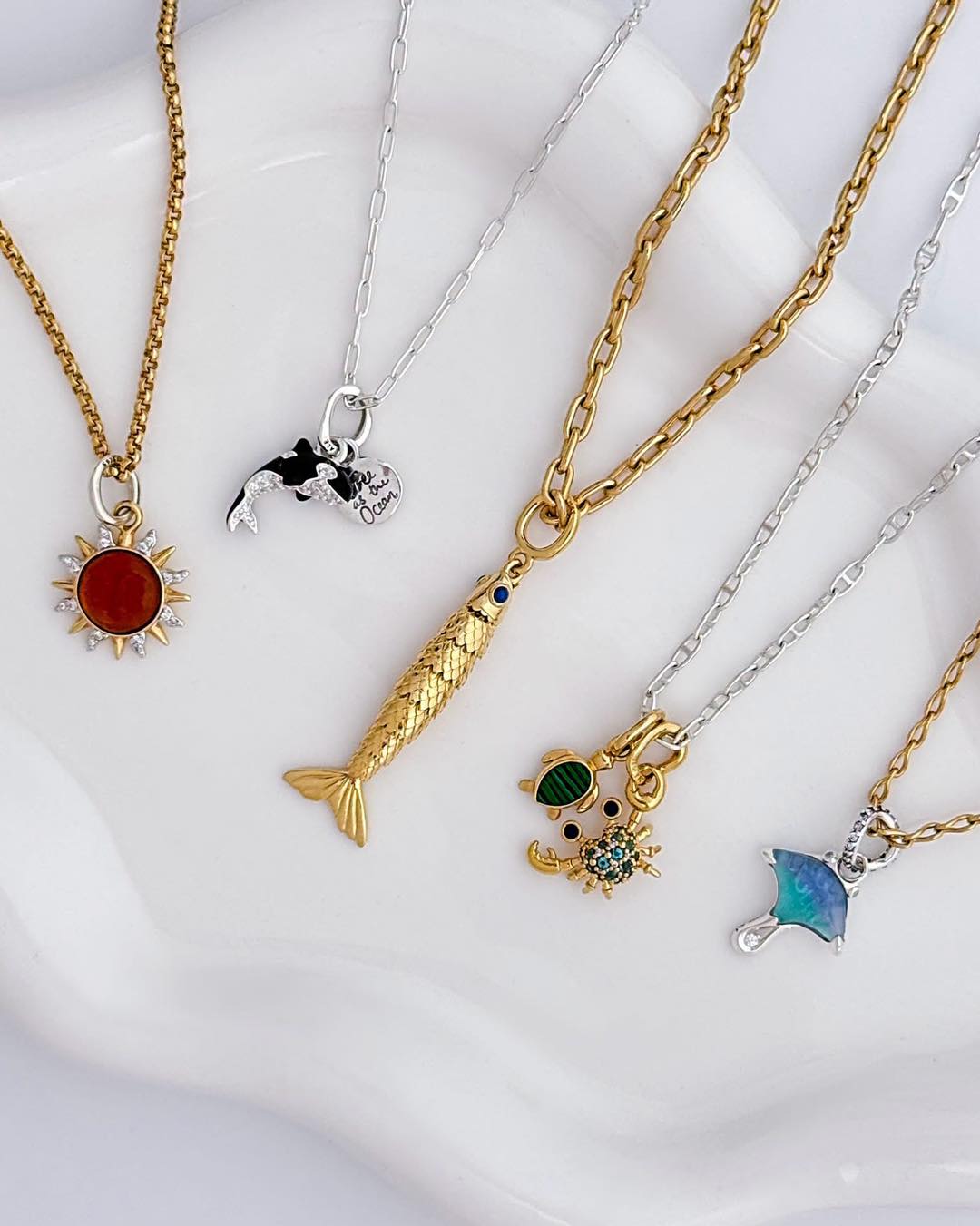 A set of five ocean-themed necklaces displayed on a white ceramic dish. The necklaces feature gold and silver chains with pendants including a red sun, a mermaid tail, a black-and-white dolphin, a sea turtle with green and blue accents, and a blue umbrella.