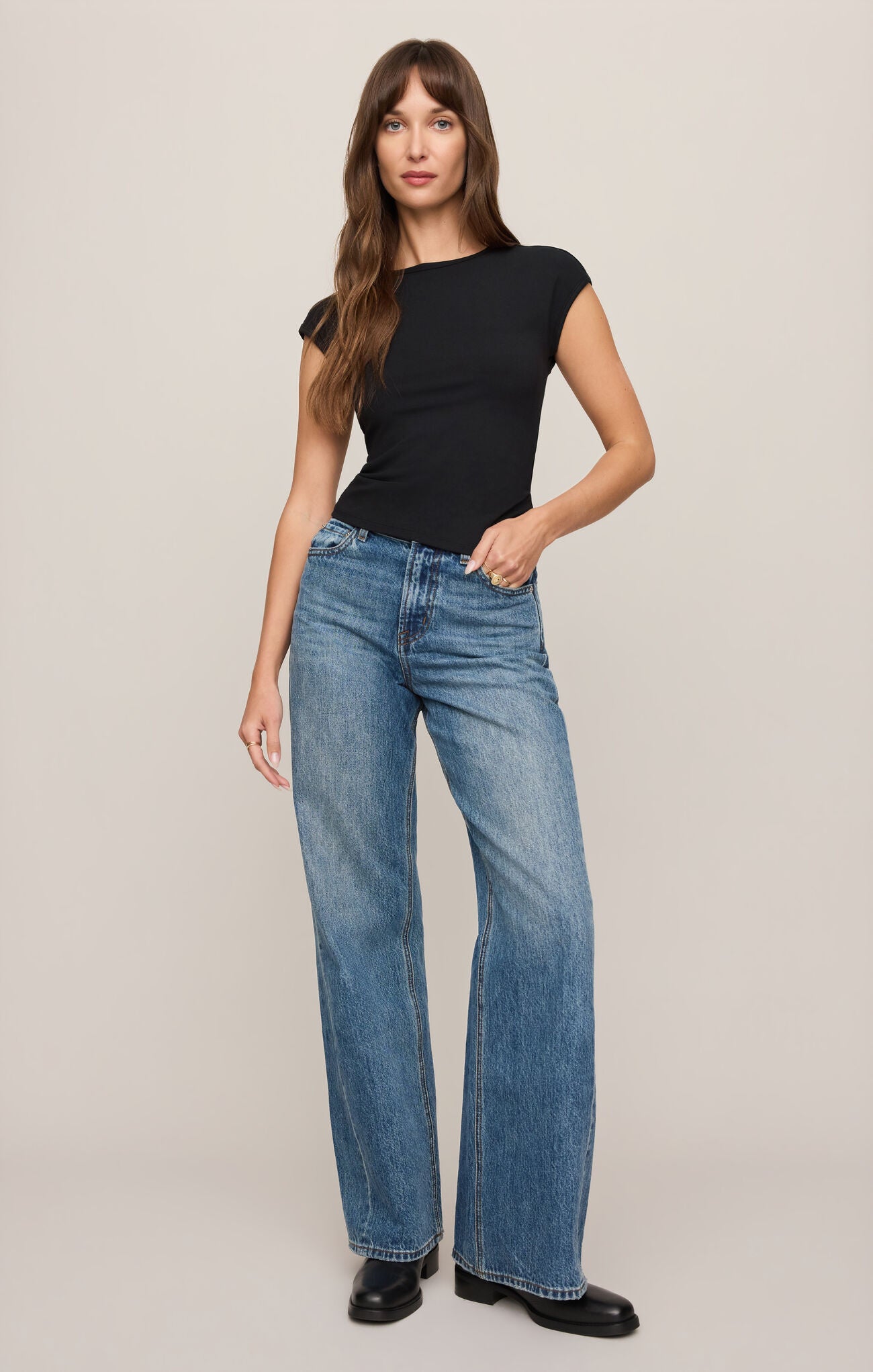 A woman modeling a black short-sleeve top and wide-leg blue jeans against a plain beige studio background.