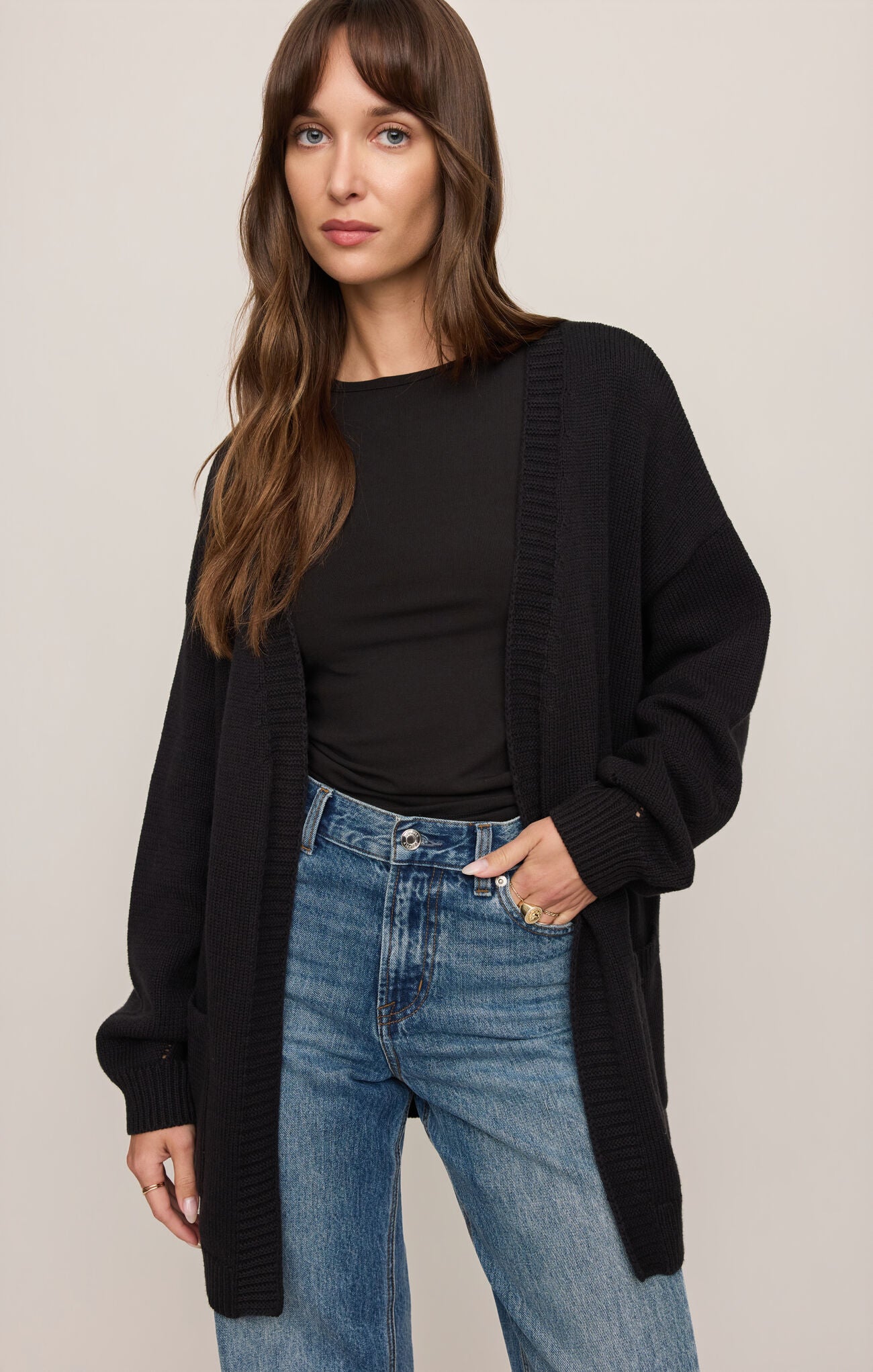 Woman wearing a black cardigan, black top, and blue jeans on a beige background