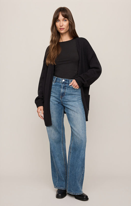 Woman wearing a black cardigan, black top, and blue jeans on a plain background