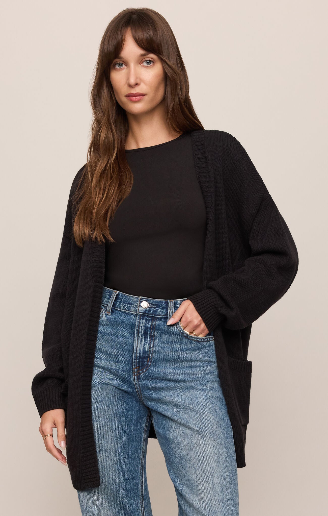 Woman wearing a black cardigan, black top, and blue jeans on a beige background