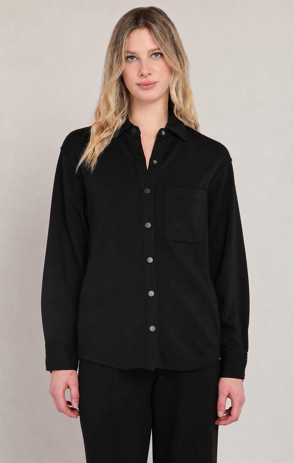 A woman modeling a black long-sleeve button-up shirt with a chest pocket, paired with black pants, against a neutral light gray studio background.