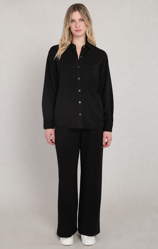 A woman modeling a black long-sleeve button-up shirt and matching black wide-leg trousers against a neutral light gray studio background.