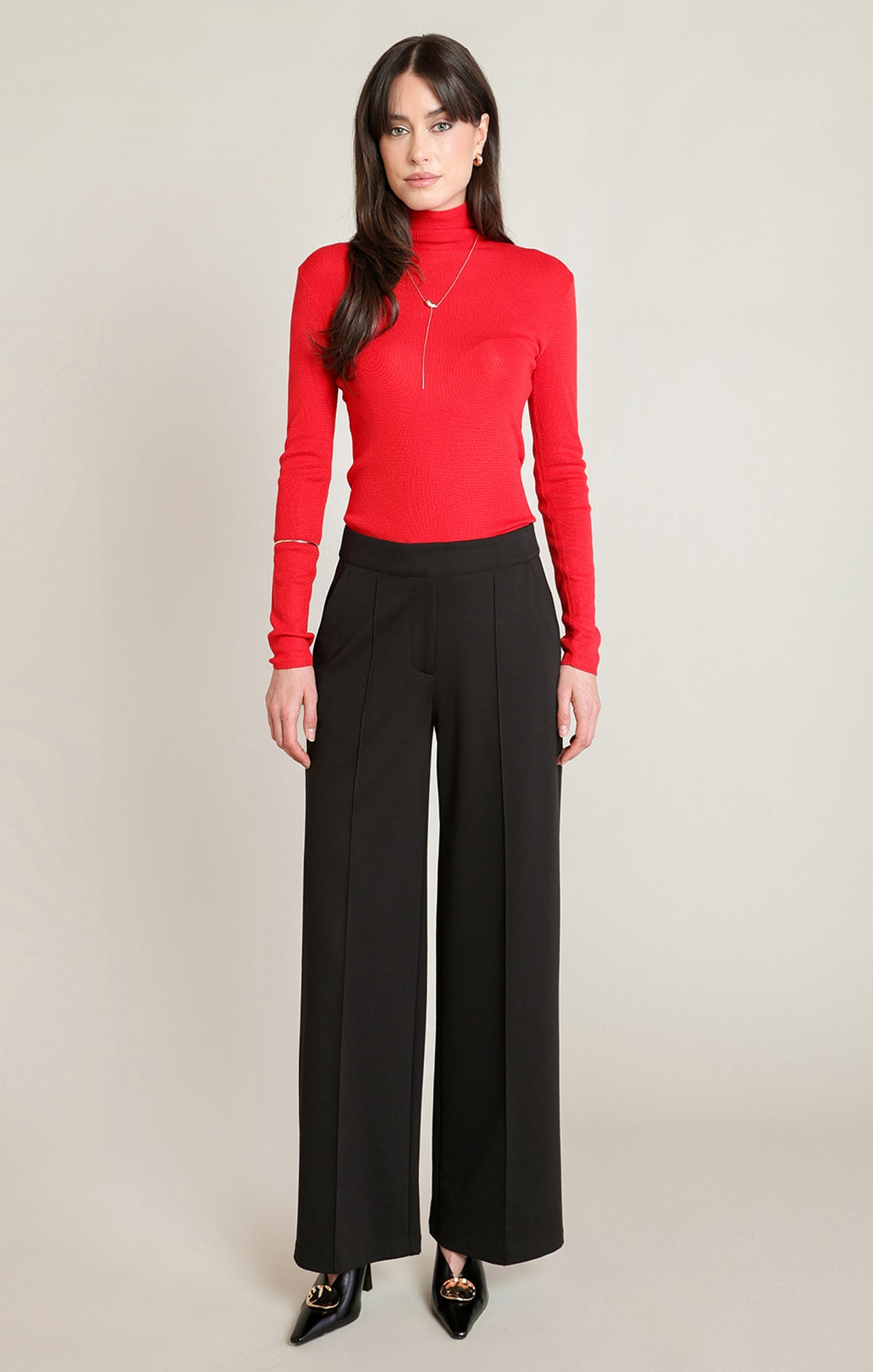 A woman modeling a bright red long-sleeve mock neck top paired with black wide-leg trousers and black pointed-toe heels, standing against a plain light beige studio background.