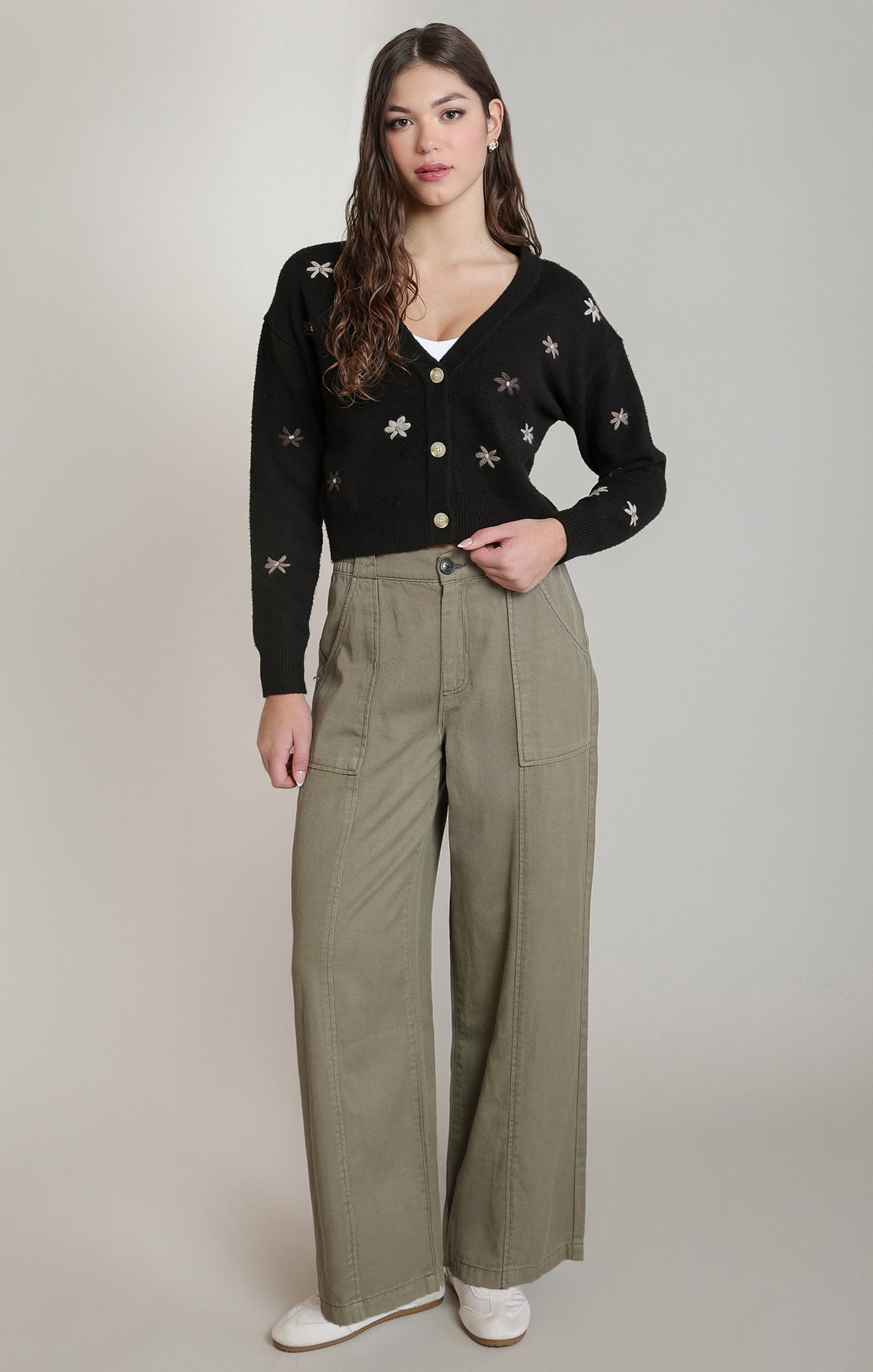 A woman modeling a black long-sleeve knit cardigan with embroidered floral details, paired with high-waisted olive green wide-leg trousers and white sneakers, against a neutral studio background.