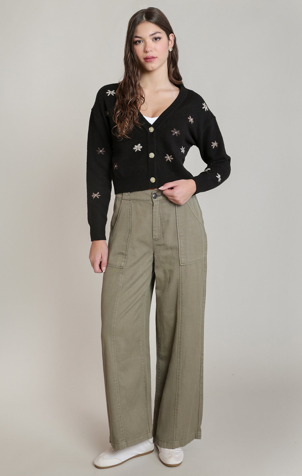 A woman modeling a black button-front cardigan with small embroidered motifs and wide-leg olive green trousers, standing against a plain light gray studio background.