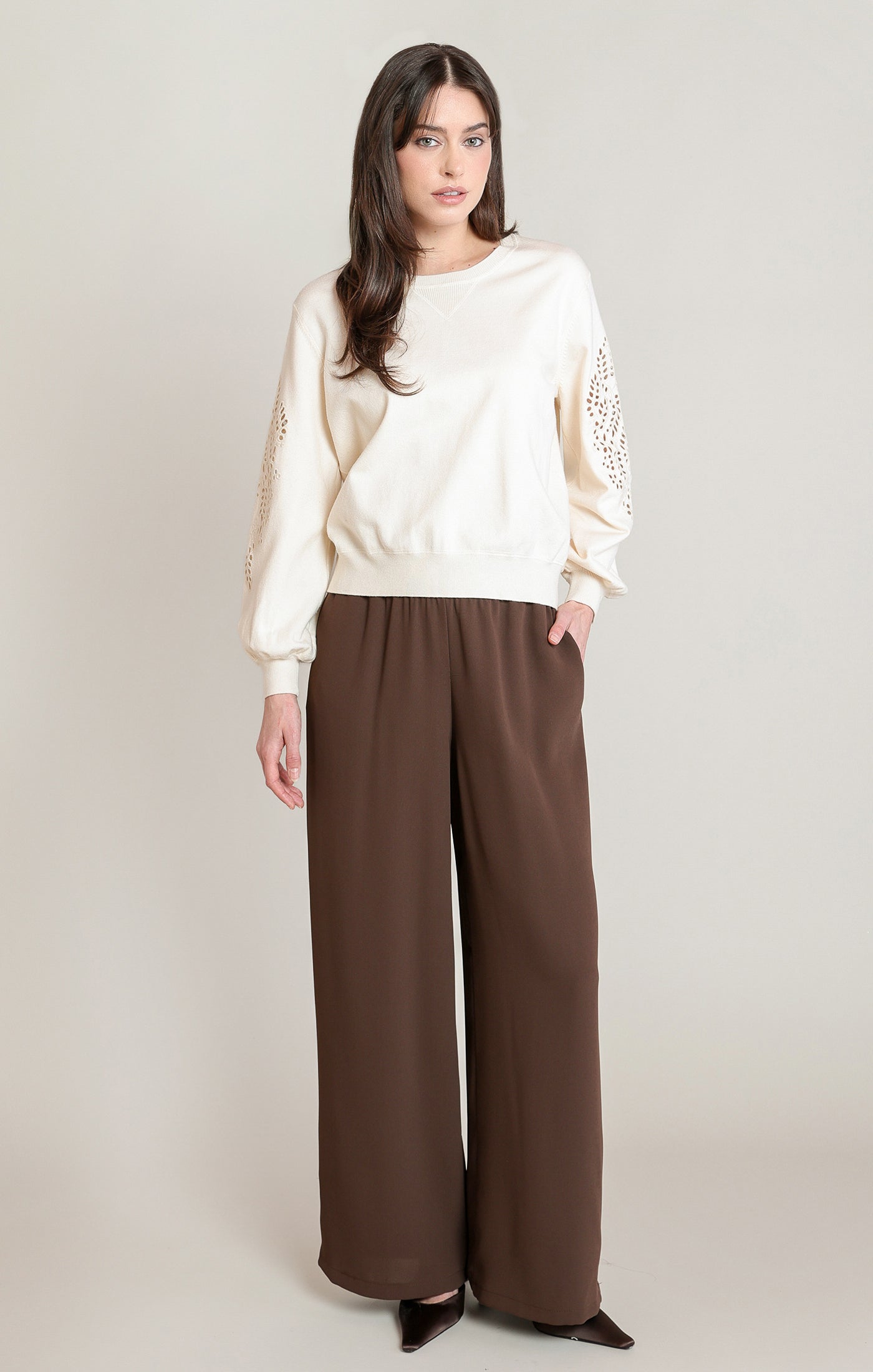 A woman modeling a cream long-sleeve top with textured sleeve details and wide-leg brown pants, standing against a plain light beige studio background.