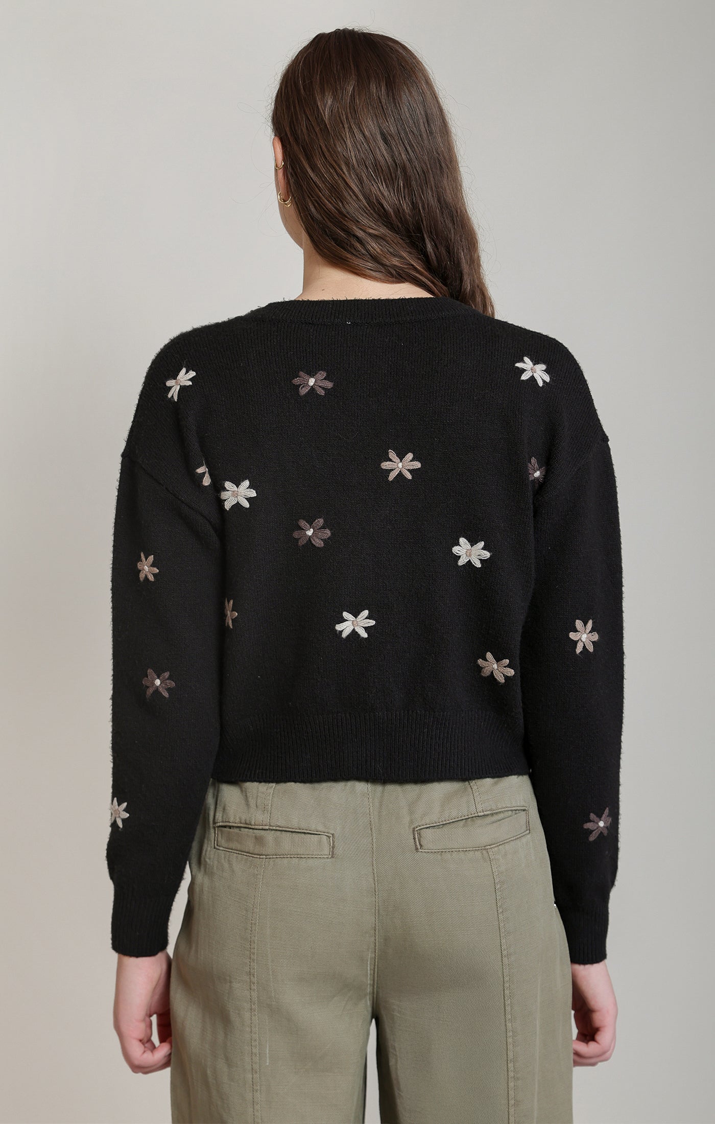 A woman viewed from the back wearing a black knit sweater with small embroidered floral motifs in light beige and gray, paired with olive green pants, against a plain light gray studio background.