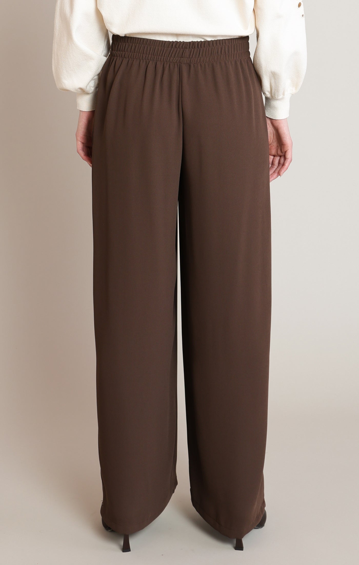 A person shown from the back wearing loose, wide-leg brown pants with an elastic waistband, paired with a white blouse and black heels, against a plain beige studio background.