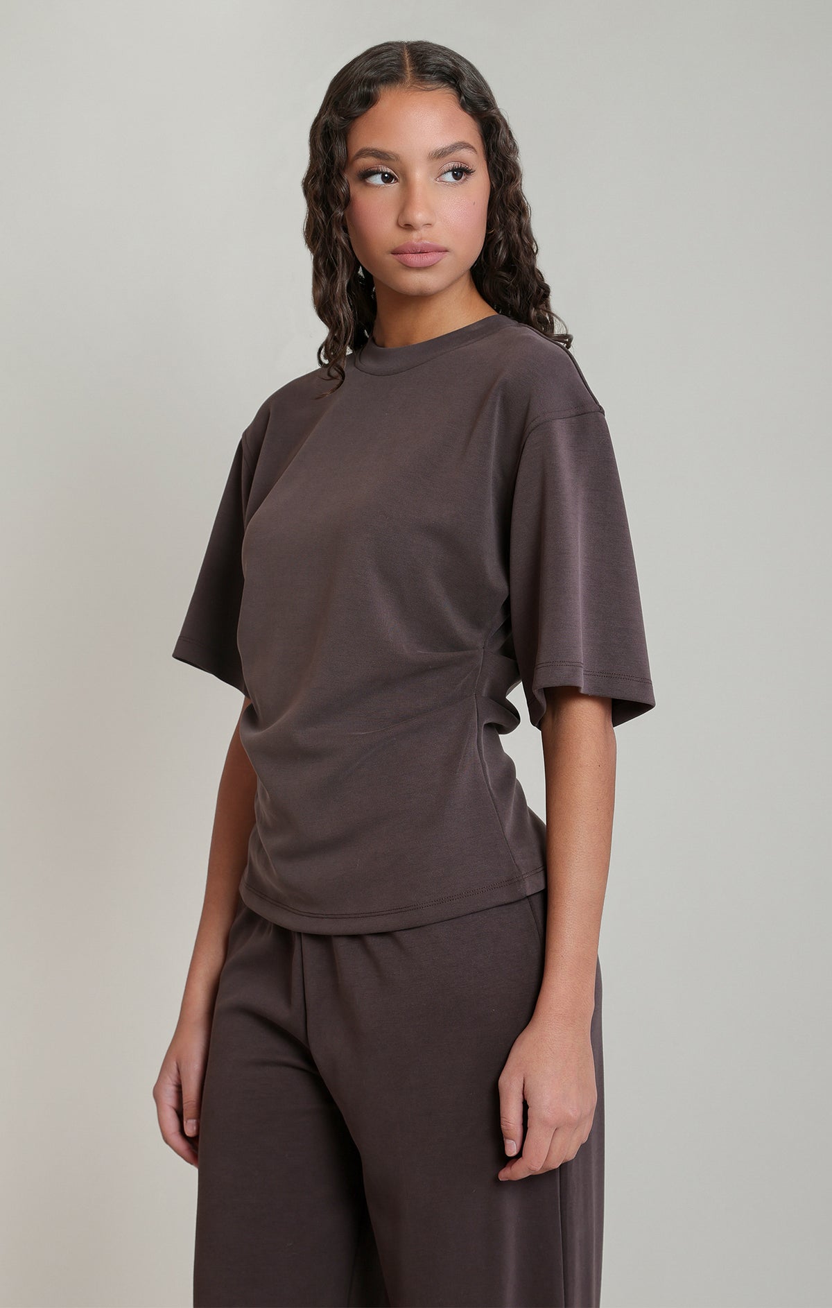 A woman modeling a dark brown short-sleeve t-shirt and matching dark brown pants against a solid light gray background.