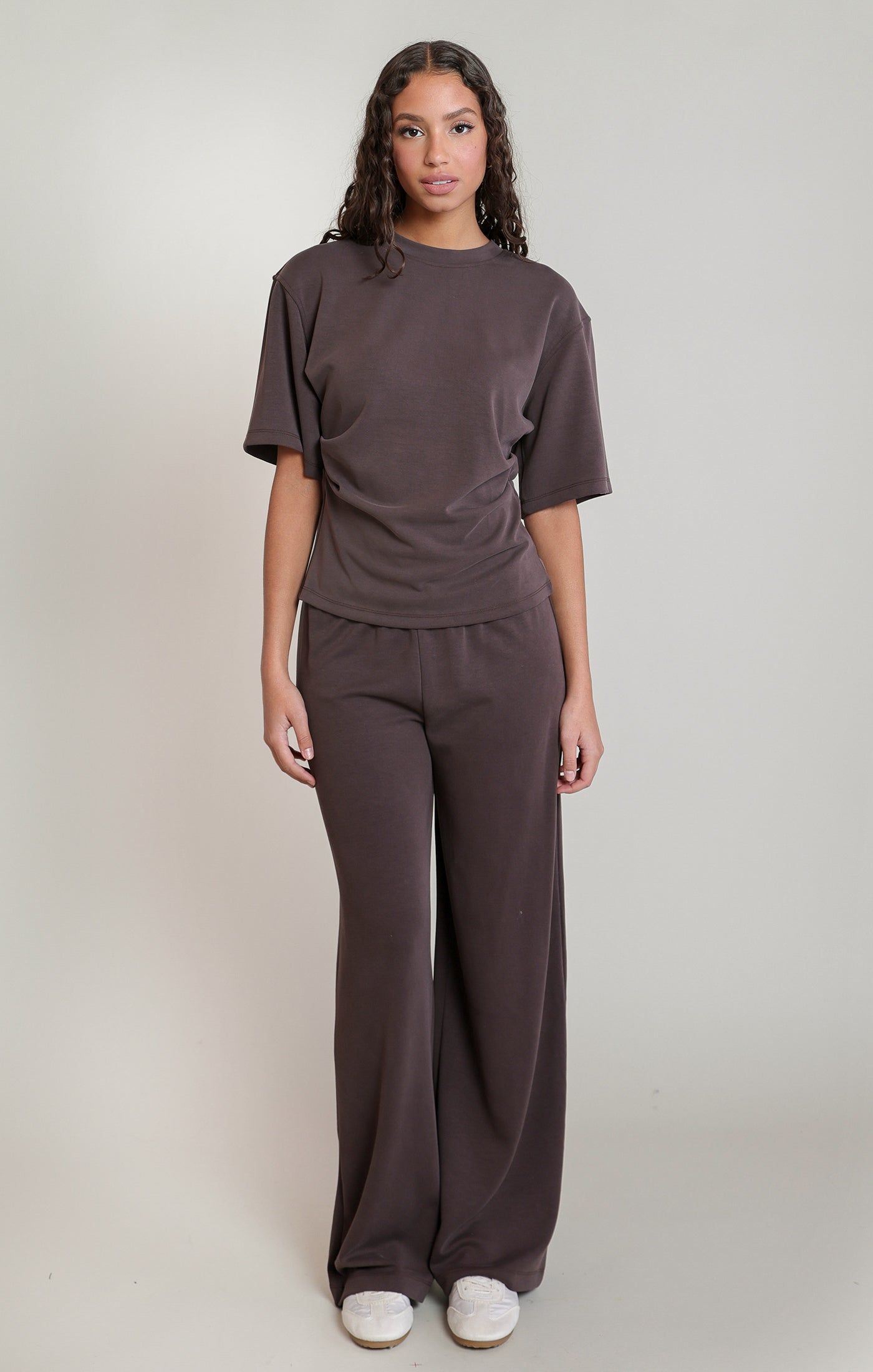 A woman modeling a matching dark brown lounge set consisting of a short-sleeve crewneck t-shirt and wide-leg pants, paired with white sneakers, against a plain light gray studio background.