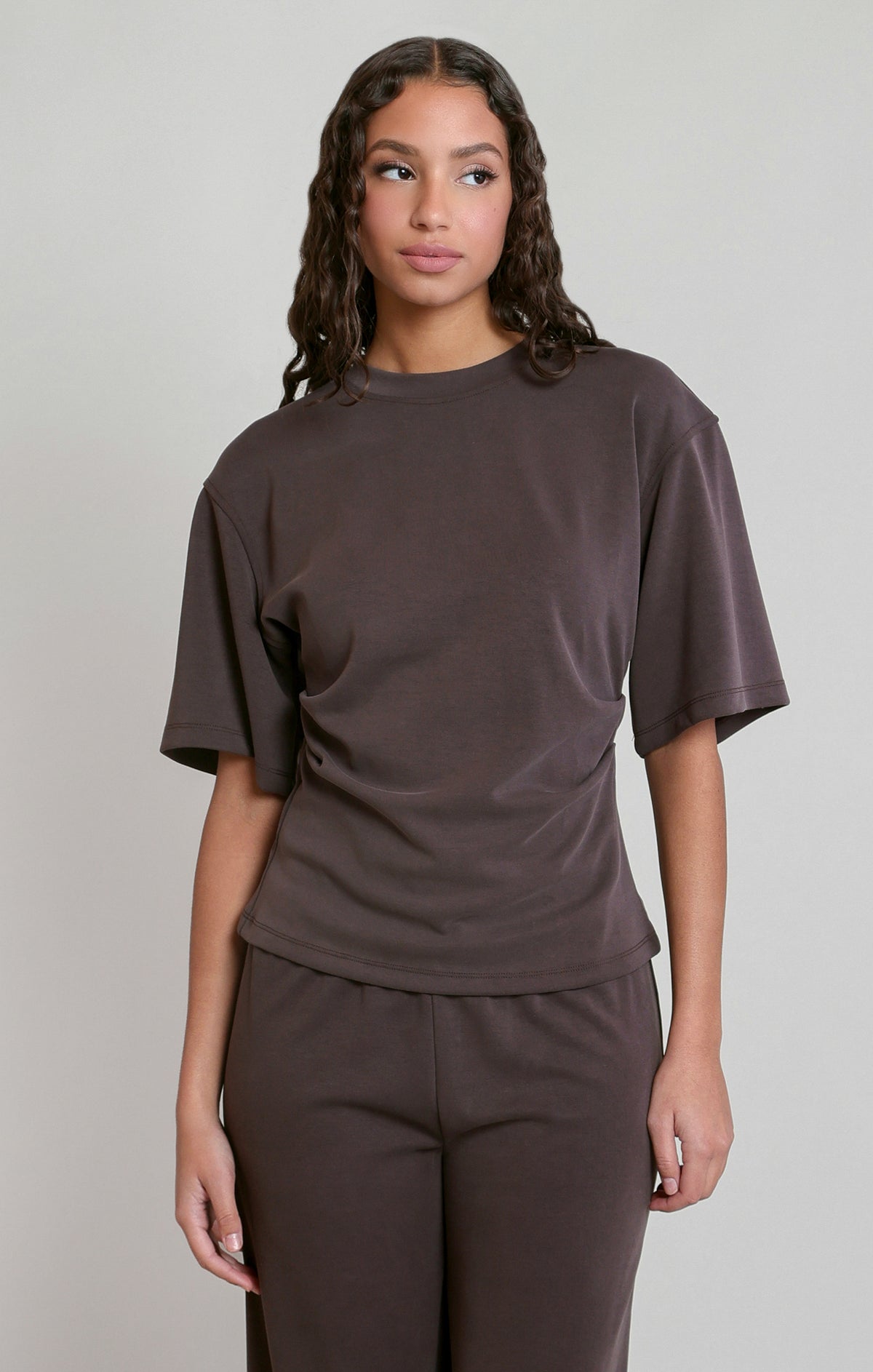 Woman wearing a brown t-shirt and pants against a plain background